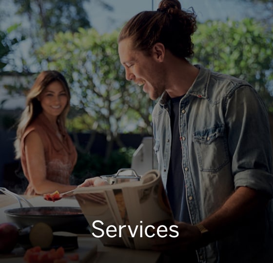 Male happily cooking dinner while holding a curled up recipe book in hand. Female smiling in background. Text over image - Services. 