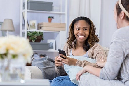 A lady with a smartphone talking to her friend sitting on a couch.