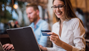 A woman wearing glasses holding a credit card whilst working on a laptop