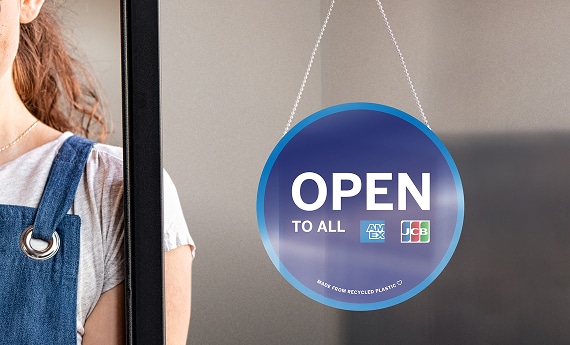 Business owner standing next to storefront with a sign that reads OPEN TO ALL and includes the AMEX logo