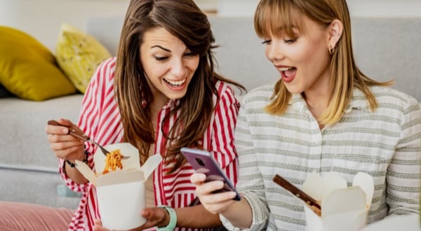 Two women eating takeout and smiling while looking at Amex App on mobile phone