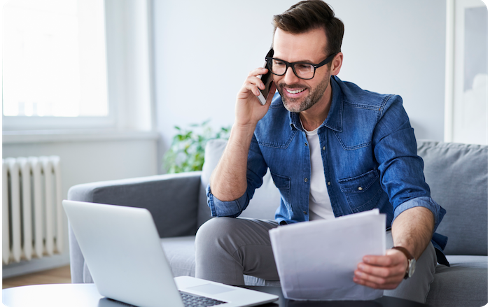 A man sitting on his couch while talking on the phone and looking at papers in his hand with laptop open