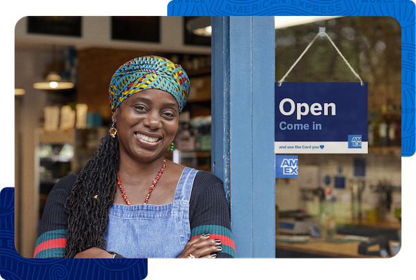A Black smiling business owner in casual clothes standing next to a sign that says “Open. Come in and use the Card you love. American Express”