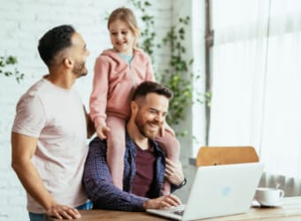 Child sitting on father's shoulders with partner looking at laptop