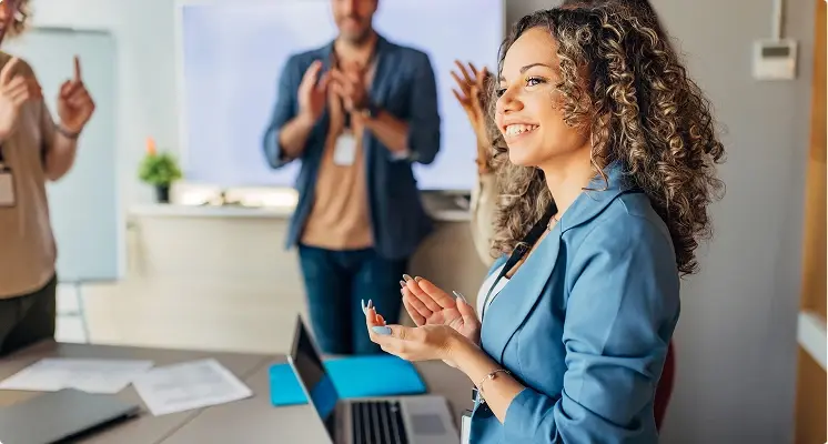 Eine junge Frau steht in einem Büro und lächelt, während sie klatscht. Im Hintergrund applaudieren Kolleginnen und Kollegen. Auf dem Tisch vor ihr steht ein Laptop.