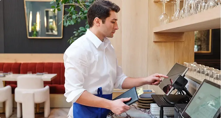 Ein Mann in weißem Hemd arbeitet in einem Restaurant an der Kasse oder einem Zahlungsterminal. In der Hand hält er ein Portemonnaie mit einer Kreditkarte.