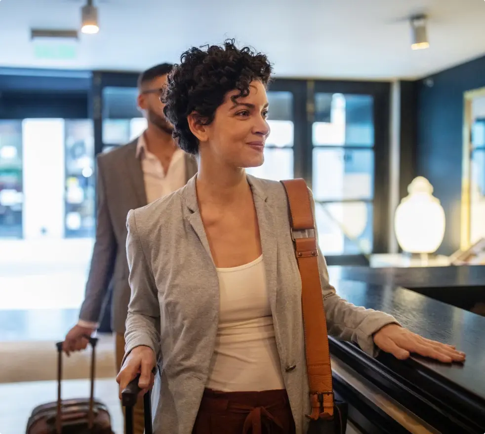 Eine Frau mit grauem Blazer checkt an einer Hotelrezeption ein. Sie hält einen Rollkoffer und lächelt, während ein Mann mit Gepäck im Hintergrund zu sehen ist.