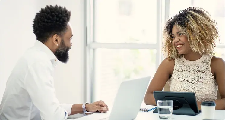 Zwei Personen sitzen sich in einem hellen Büro gegenüber, lächeln und unterhalten sich. Auf dem Tisch stehen ein Laptop, ein Tablet und ein Glas Wasser.