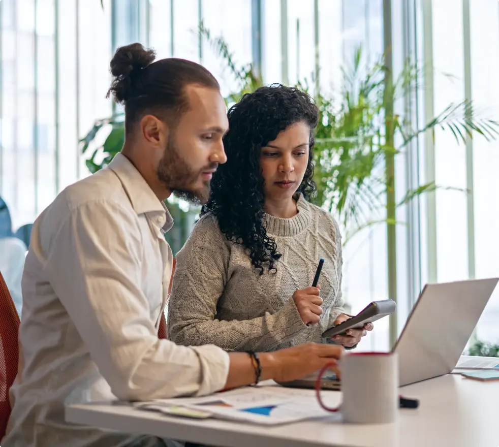 Zwei Personen sitzen in einem modernen Büro an einem Schreibtisch, arbeiten gemeinsam an einem Laptop und besprechen Unterlagen. Im Hintergrund sind große Fenster und grüne Pflanzen zu sehen.
