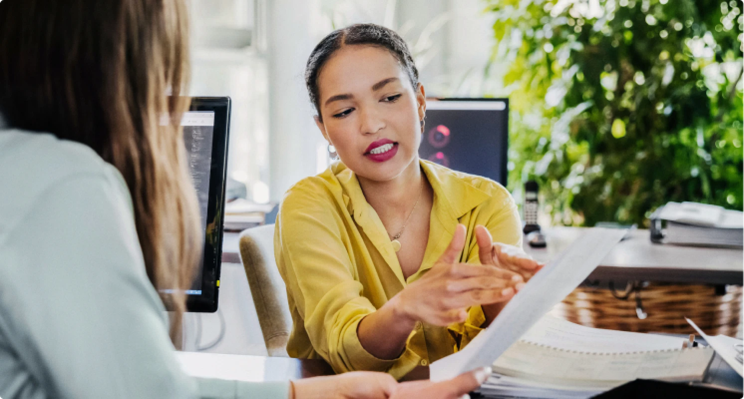 Zwei Personen sitzen in einem modernen Büro an einem Schreibtisch. Eine Frau in gelber Bluse erklärt einer Kollegin ein Dokument und zeigt auf bestimmte Stellen darauf.