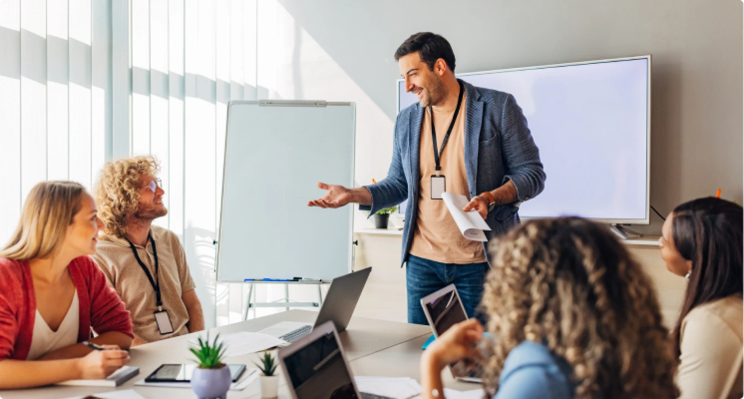 Ein Mann steht in einem Konferenzraum vor einem Whiteboard und spricht zu seinem Team. Mehrere Kolleginnen und Kollegen sitzen am Tisch und hören aufmerksam zu.