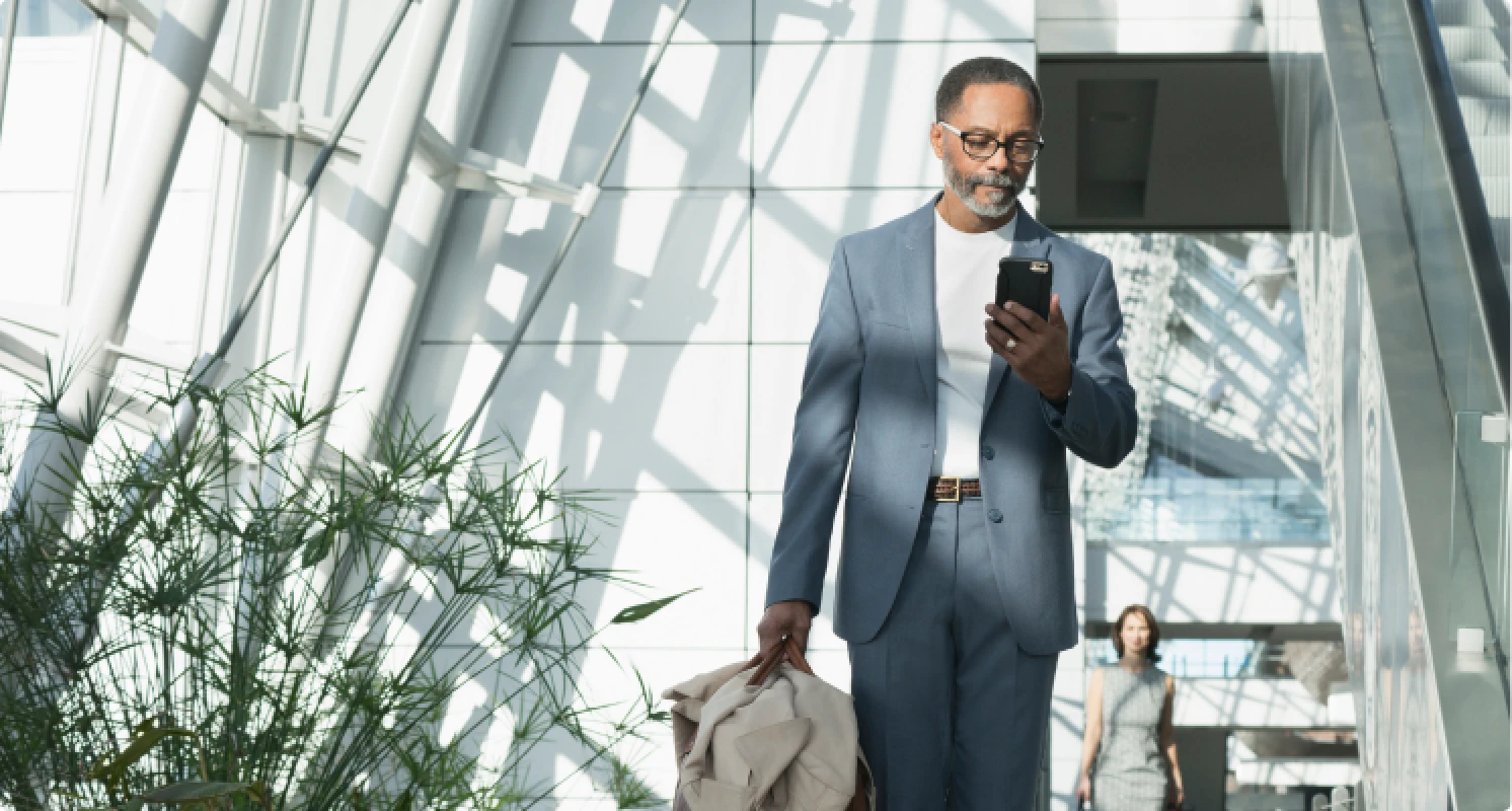 Ein Mann in hellblauem Anzug läuft durch ein lichtdurchflutetes Gebäude und schaut auf sein Smartphone. In der anderen Hand hält er eine Jacke.