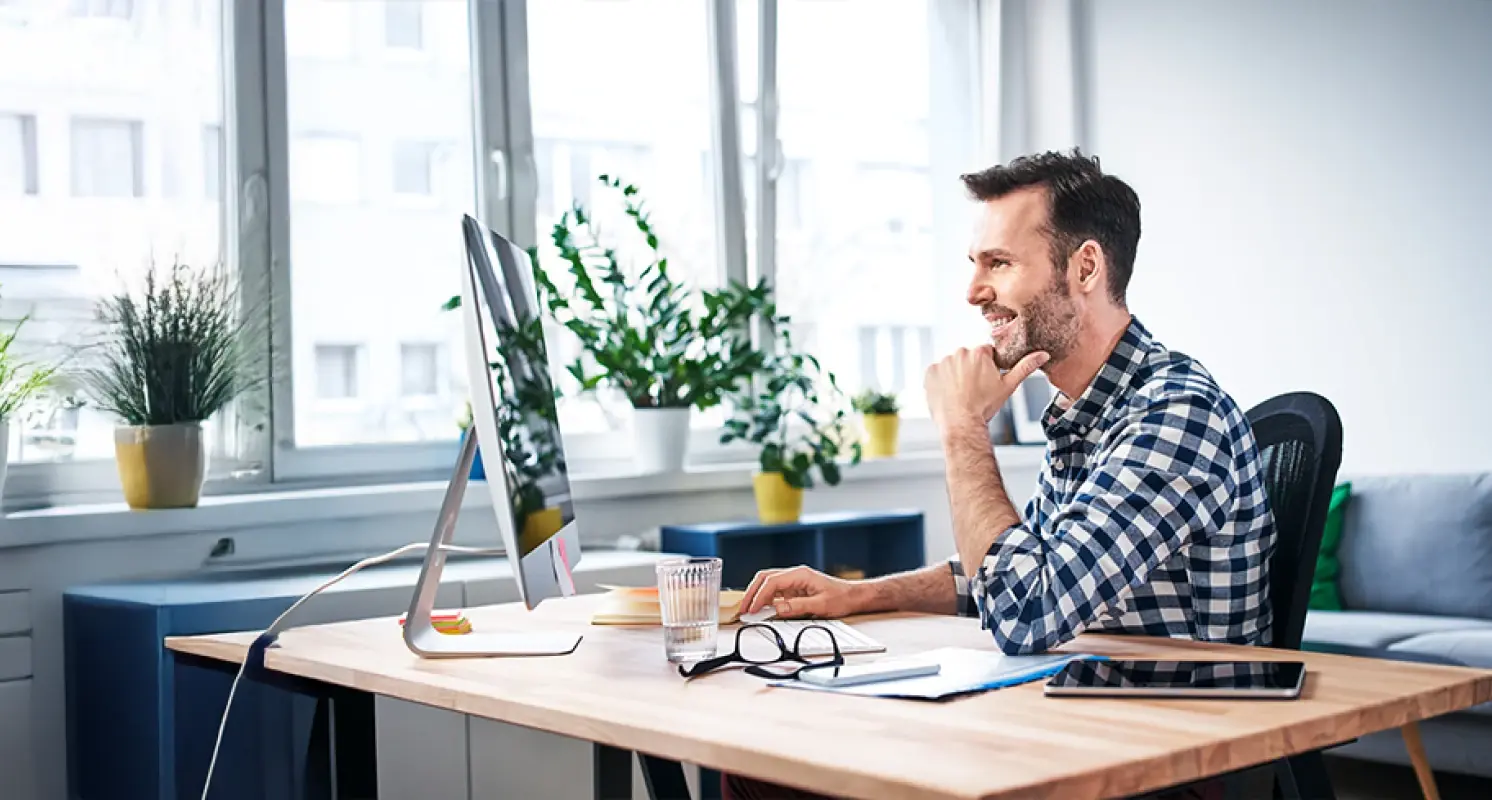 Mann arbeitet lächelnd am Computer in einem hellen Büro mit vielen Pflanzen am Fenster.