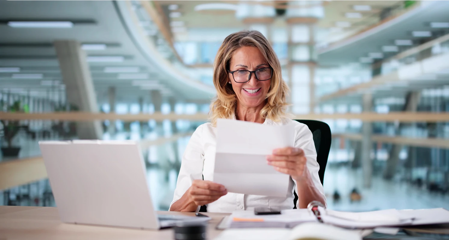 Eine lächelnde Frau mit Brille sitzt in einem modernen Büro an einem Schreibtisch, hält ein Dokument in der Hand und liest es zufrieden durch.
