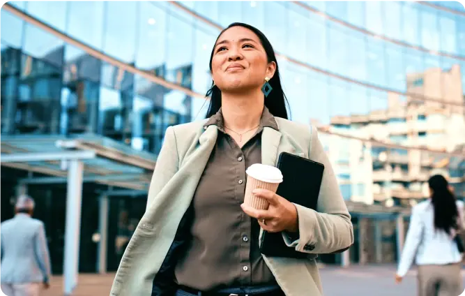 Geschäftsfrau geht selbstbewusst mit Kaffeebecher und Tablet vor einem modernen Bürogebäude.