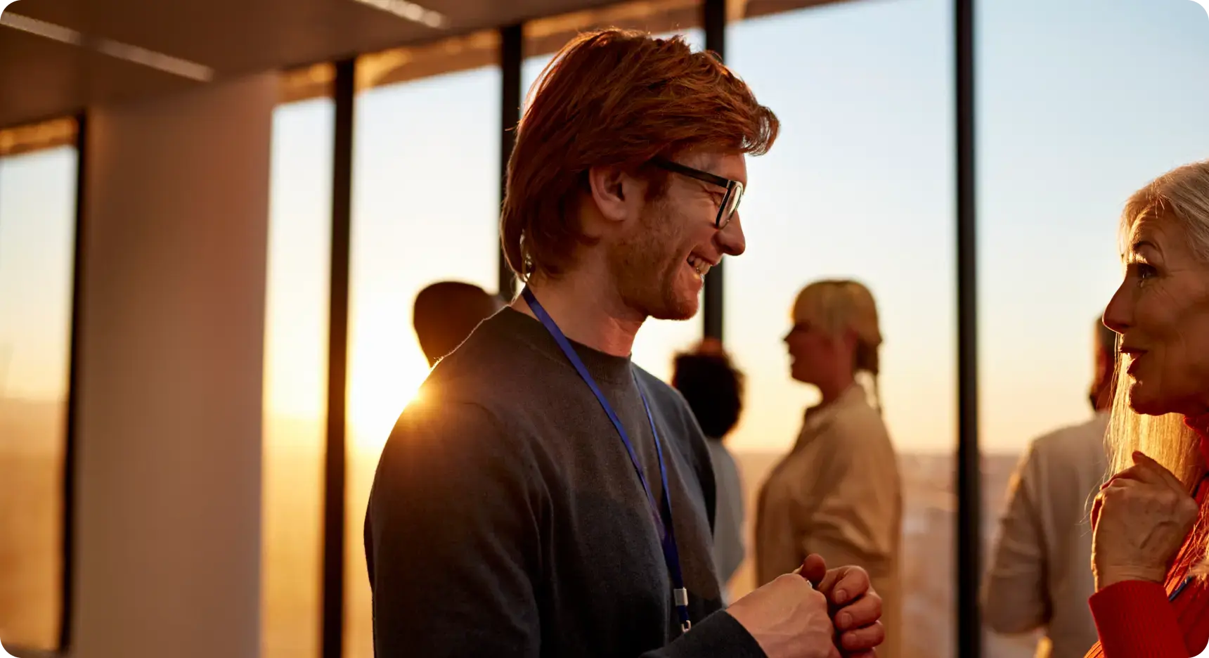 Zwei Personen interhalten sich lächelnd bei iinem Networking-Event in einem modernen Innenraum vor großen Fenstern bei Sonnenuntergang.