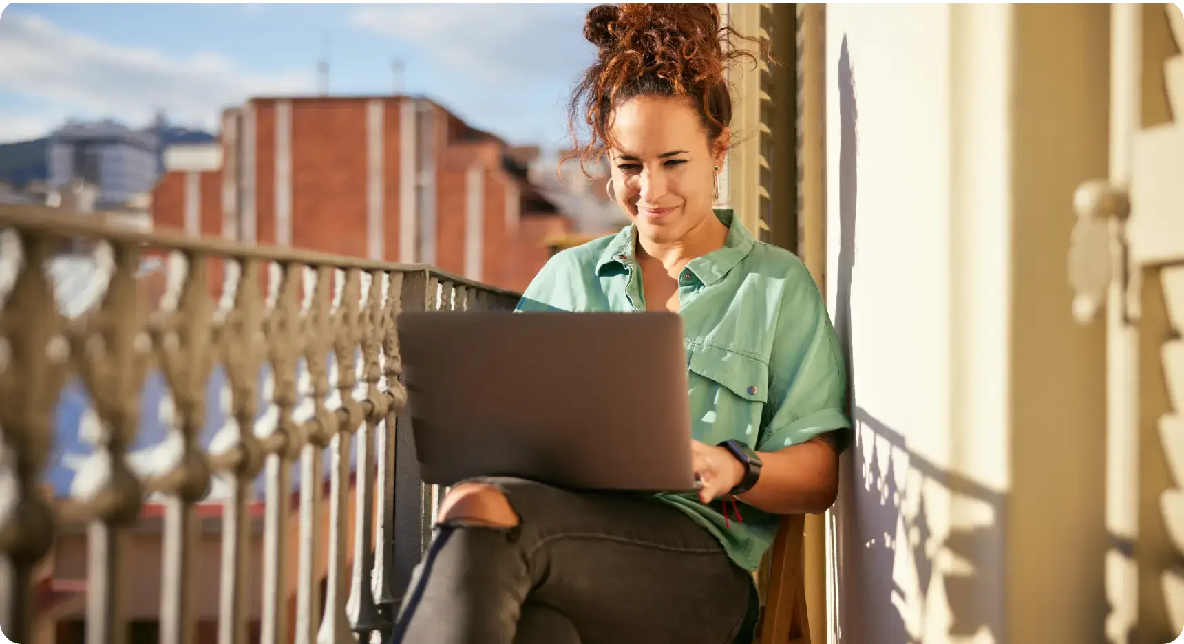 Frau sitzt auf einem Balkon in der Sonne und arbeitet entspannt an einem Laptop.