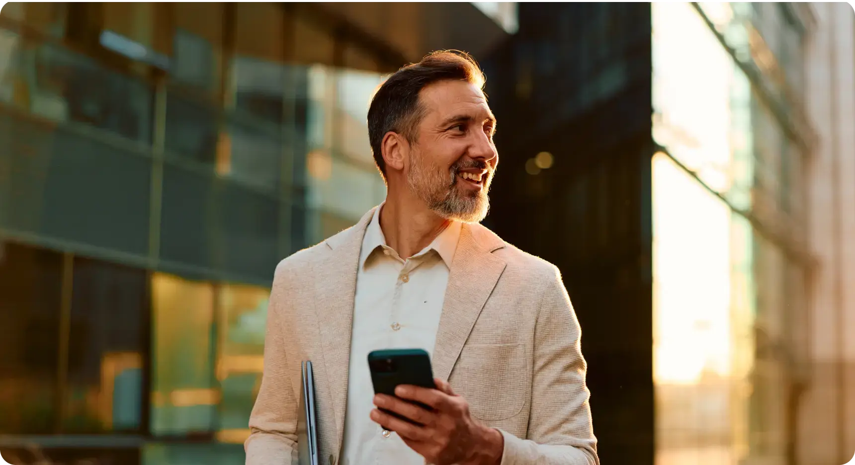 Lächelnder Geschäftsmann läuft draußen an modernen Bürogebäuden vorbei und schaut auf sein Smartphone.