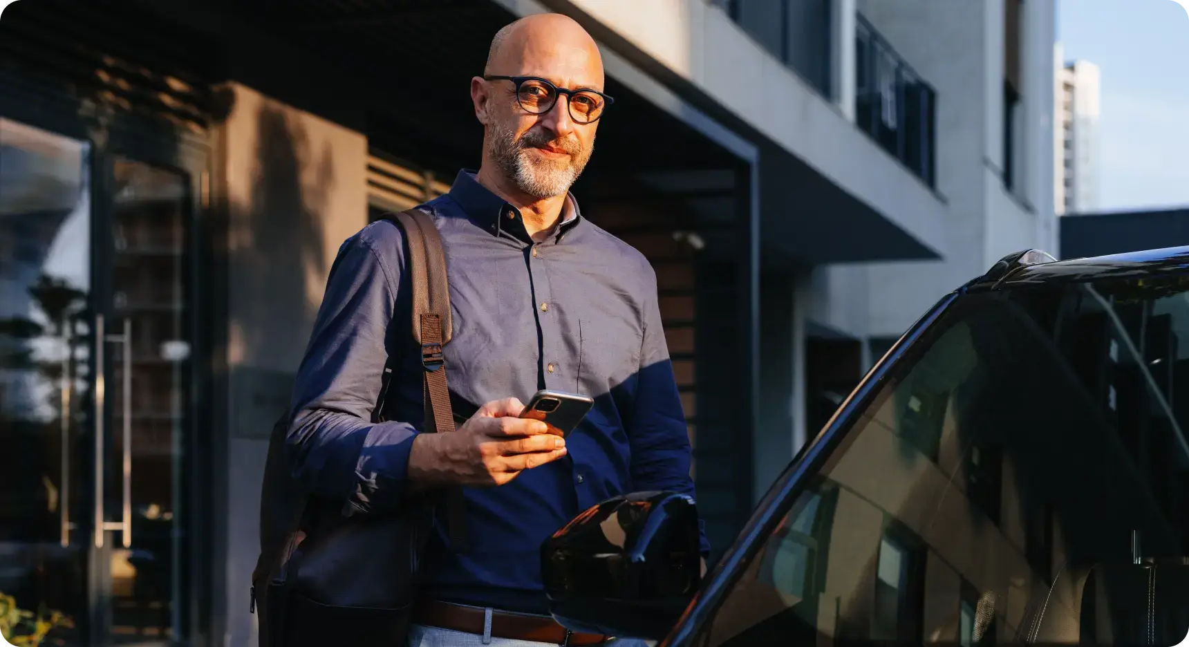 Geschäftsmann mit Brille steht neben einem Auto und blickt auf sein Smartphone.