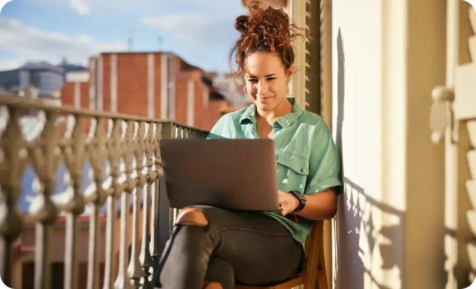 Frau sitzt auf einem Balkon in der Sonne und arbeitet entspannt an einem Laptop.
