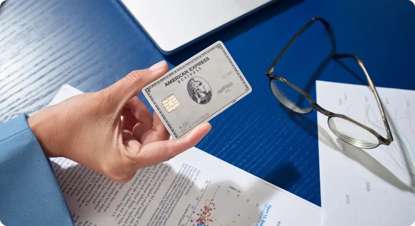Hand hält eine American Express Business Platinum Card über Geschäftsdokumenten, daneben liegen eine Brille und ein Laptop.