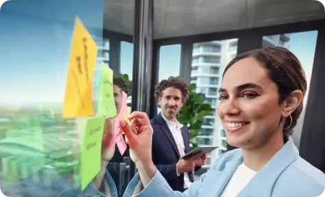 Geschäftsfrau schreibt Ideen auf bunte Post-its an einer Glaswand, während ein Kollege im Hintergrund Notizen auf einem Tablet macht.