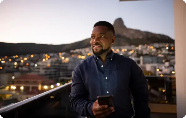 Mann steht auf einem Balkon mit Blick über eine Stadt und hält ein Smartphone
