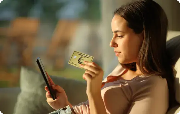 Frau sitzt auf einem Sofa und hält eine American Express Business Gold Card und ein Smartphone