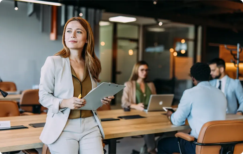 rau im Business-Outfit mit Klemmbrett steht selbstbewusst in einem modernen Konferenzraum, im Hintergrund arbeiten Kollegen an Laptops.