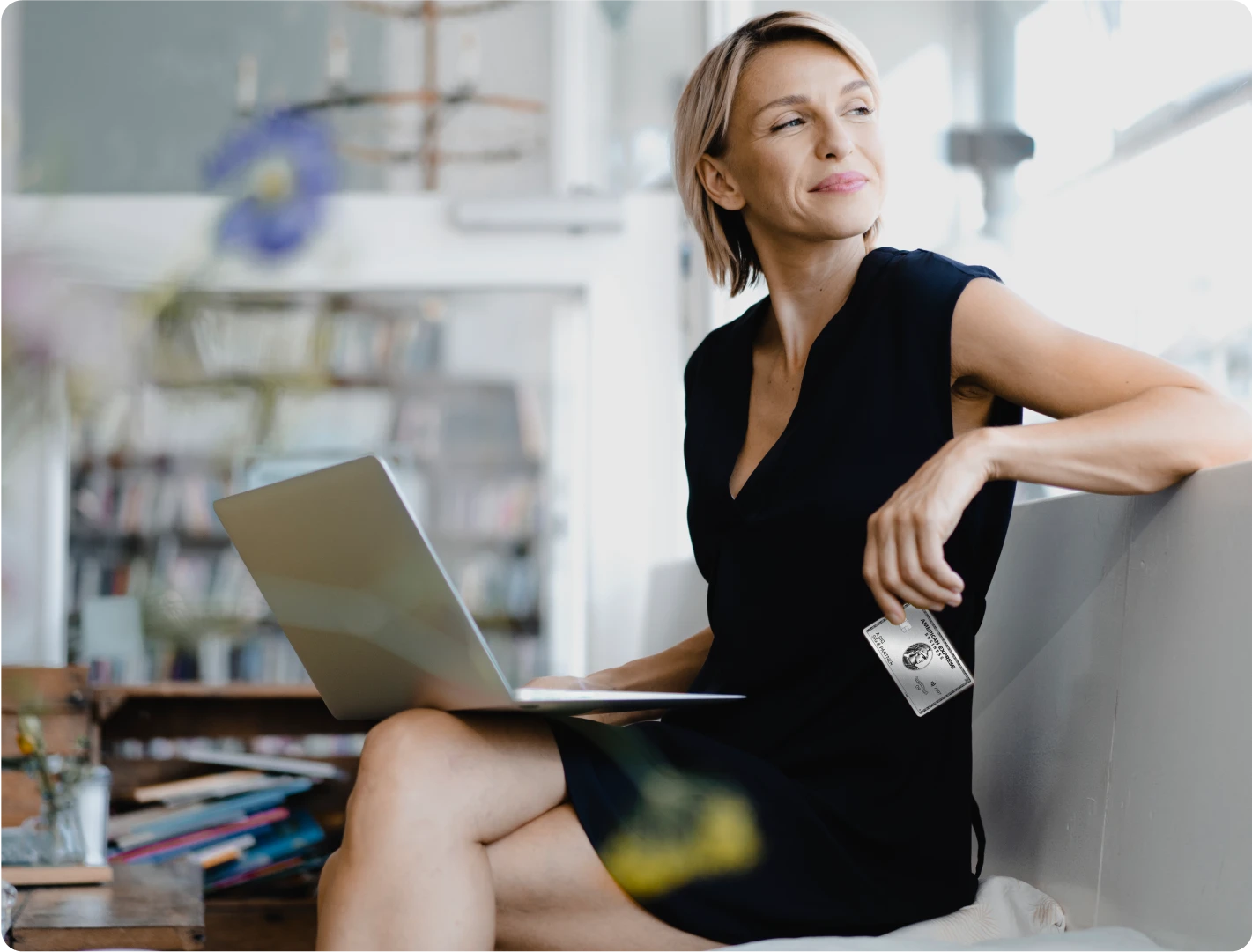 Frau sitzt entspannt mit Laptop in einem Café, hält eine American Express Business Platinum Card in der Hand und schaut lächelnd zur Seite.