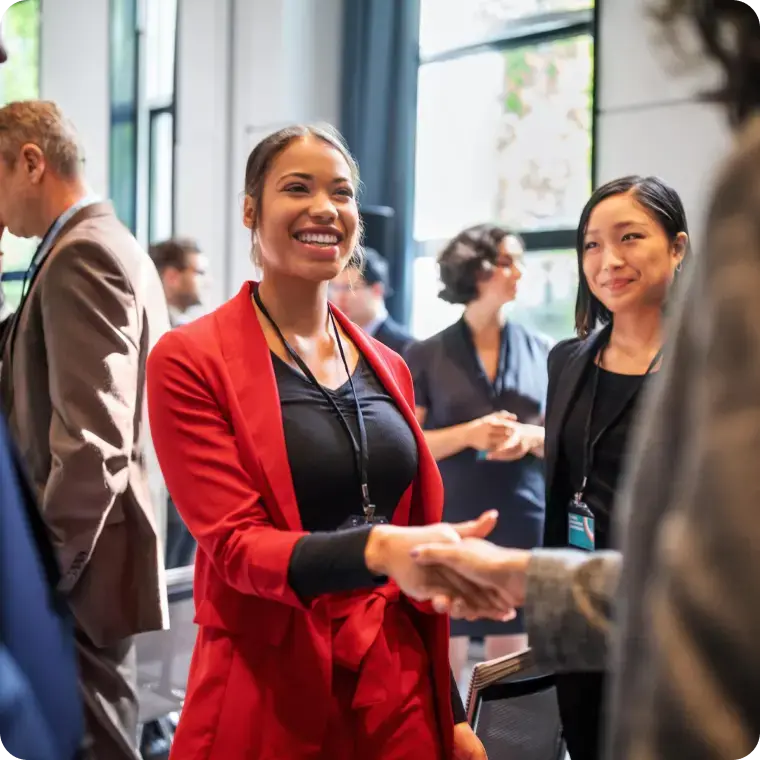 Frau in rotem Blazer schüttelt auf einer Veranstaltung einer Person die Hand.