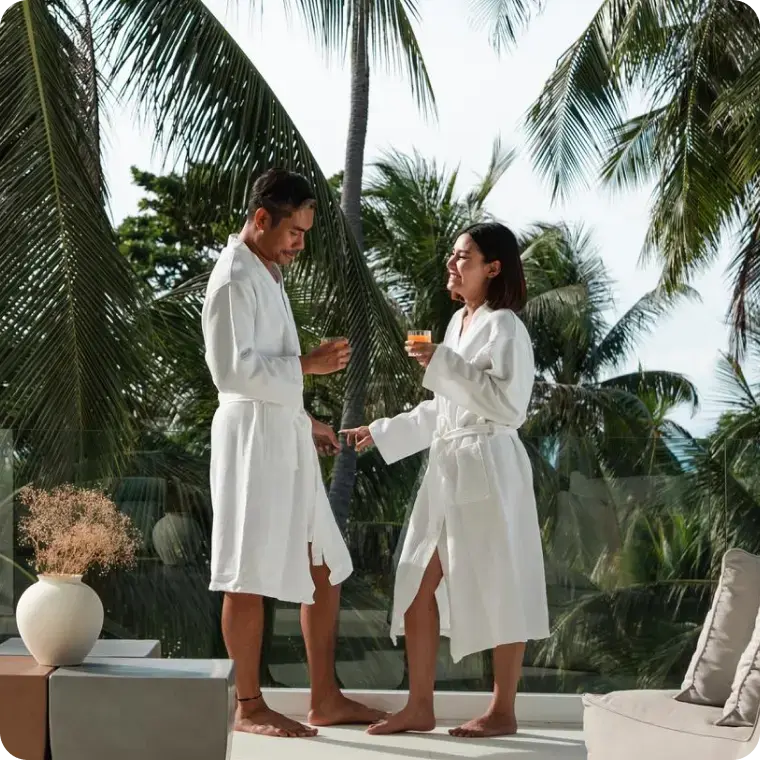 Paar in weißen Bademänteln trinkt auf einer Terrasse mit Palmen im Hintergrund.