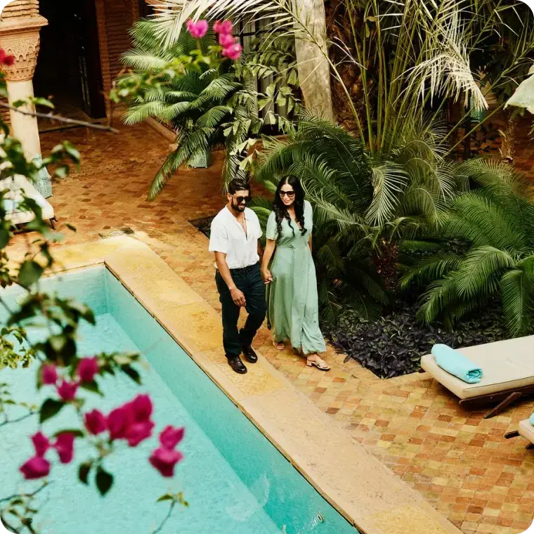 Paar geht Hand in Hand an einem Pool entlang, umgeben von tropischen Pflanzen.