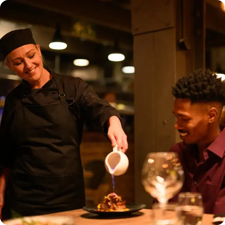 Köchin gießt Sauce über ein Gericht, während ein Gast am Tisch sitzt.