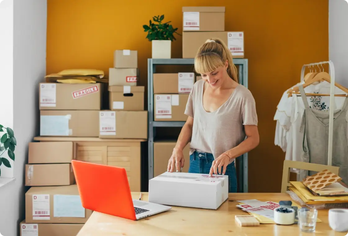 Frau verpackt Produkte in einem Raum mit Regalen voller Kartons und Versandmaterialien.