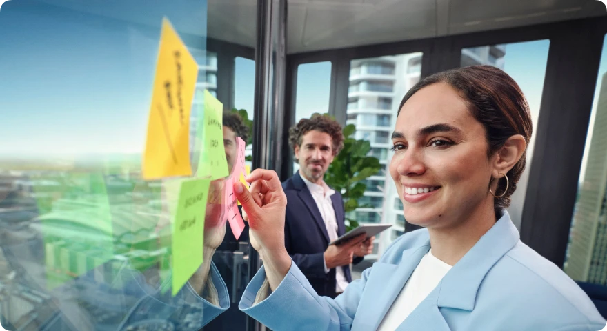 Geschäftsfrau schreibt Ideen auf bunte Post-its an eine Glaswand, während ein Kollege mit Tablet im Hintergrund steht.