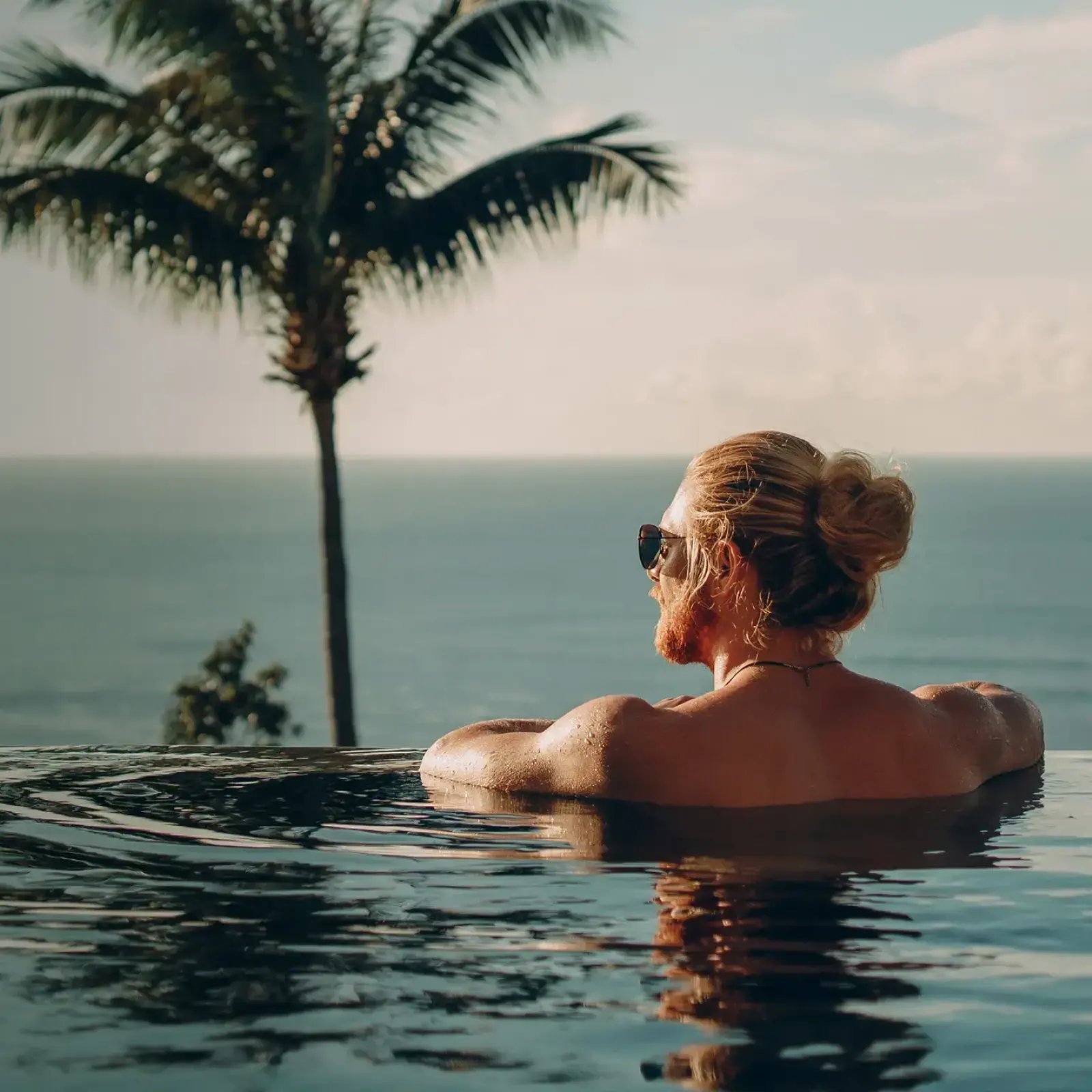 Person lehnt im Poolrand und blickt aufs Meer. Im Hintergrund steht eine Palme.