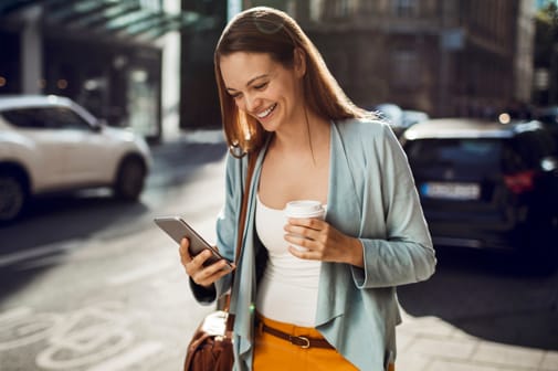 Lächelnde Frau geht bei Sonnenschein durch die Stadt, hält einen Kaffeebecher in der einen Hand und ihr Smartphone in der anderen.