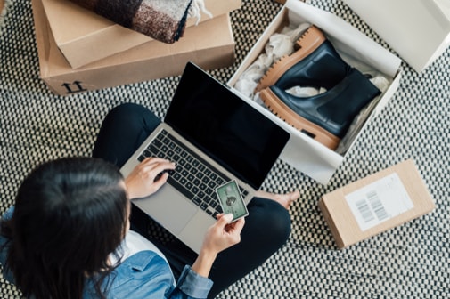 Frau sitzt auf dem Boden mit Laptop und ihrer Amex, umgeben von Paketen und einem geöffneten Schuhkarton.