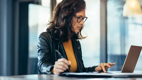  Junge Frau mit Brille und Lederjacke sitzt konzentriert an einem Laptop und schreibt etwas auf Papier in einem modernen Büro.