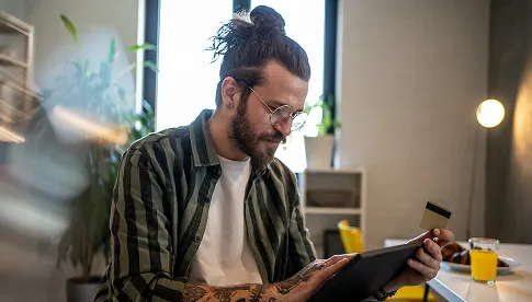  Mann mit Brille, Dutt und tätowierten Armen sitzt am Tisch, hält ein Tablet und eine Kreditkarte in der Hand, im Hintergrund steht ein Glas Orangensaft.