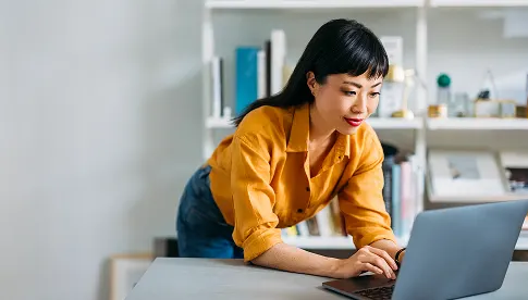  Frau in gelbem Hemd lehnt sich über einen Tisch und arbeitet konzentriert an einem Laptop in einem hellen, modernen Raum.