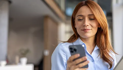 Frau mit langen roten Haaren schaut lächelnd auf ihr Smartphone, steht in einem modernen Büro.