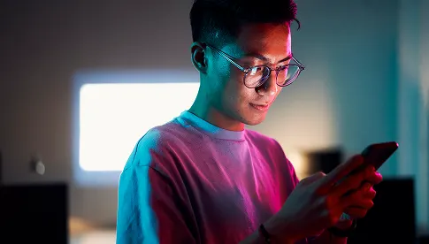  Junger Mann mit Brille und kurz geschnittenen Haaren steht in einem dunklen Raum mit violett-blauer Beleuchtung und schaut auf sein Smartphone.