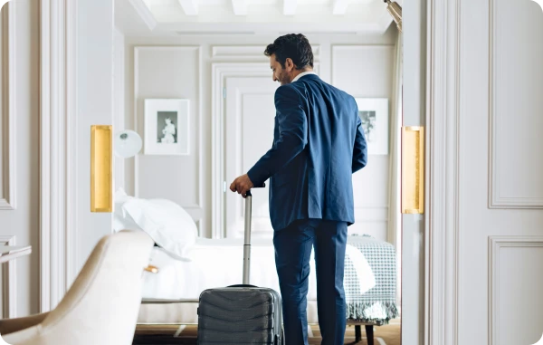 American Express Online Reisen - Mann im Hotelflur mit Koffer und über der Schulter getragenem Sakko.