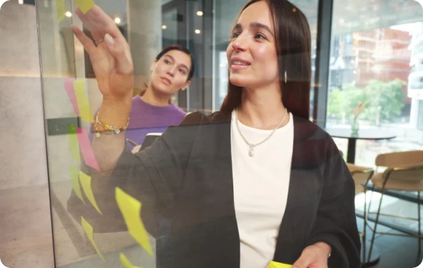 B4B Marketplace - Frau im Business-Outfit steht vor einer Glaswand mit gelben Klebezetteln, im Hintergrund Kolleg:innen beim Brainstorming.