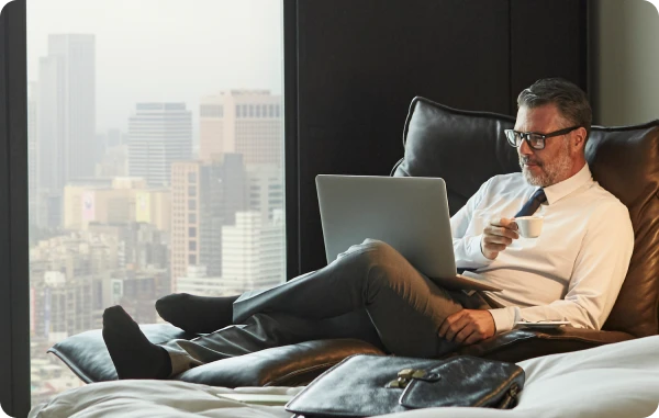 Fine Hotels & Resorts - Mann sitzt im Hotelzimmer auf dem Bett mit Blick auf eine Großstadt, arbeitet am Laptop.