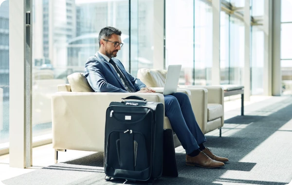 Online-Kartenkonto - Geschäftsmann sitzt mit Koffer in heller Wartehalle, schaut auf sein Smartphone.