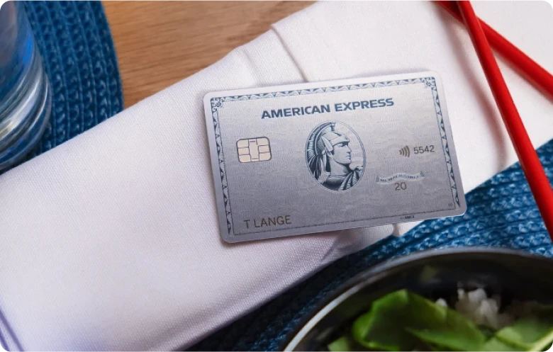 American Express Business Platinum Karte aus Metall liegt schräg auf weißer Stoffserviette neben einem Glas und roten Essstäbchen auf einem blauen Tischset.