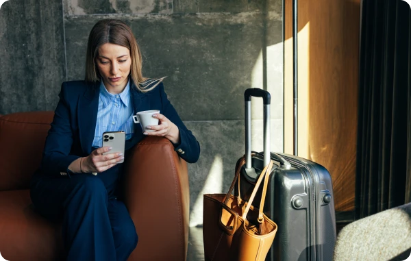 Reisegepäck-Versicherung - Frau sitzt mit Smartphone auf einem Sessel, ihr Gepäck steht neben ihr.